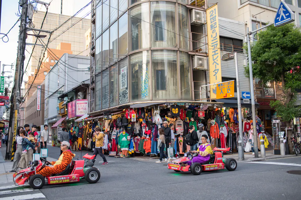 Two people in colorful go-karts dressed in costumes drive through a busy urban street with a clothing store. Pedestrians walk nearby, and the store displays a variety of vibrant outfits on hangers. Buildings and signs fill the background.