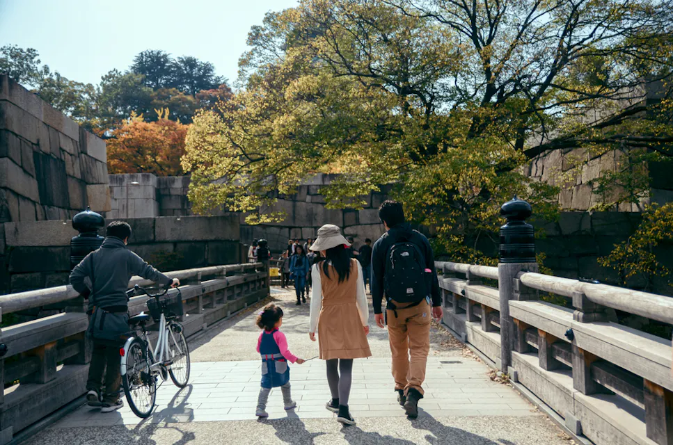 A family walks along a wooden bridge surrounded by trees in autumn. A child in a pink jacket holds hands with her parents, while a person with a bicycle stands nearby. People walk ahead, enjoying the sunny day.