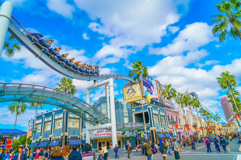 A vibrant theme park scene with a roller coaster in mid-loop, colorful buildings, and palm trees under a bright blue sky. Visitors stroll along the path, enjoying the lively atmosphere.