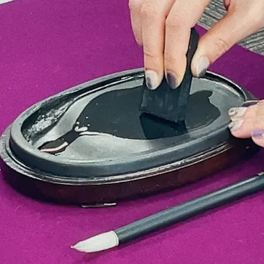Calligraphy Workshop A person uses a black ink stick on a wet inkstone to grind ink, with a pencil and a small bottle nearby, all placed on a purple surface.