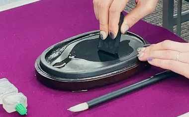 A person uses a black ink stick on a wet inkstone to grind ink, with a pencil and a small bottle nearby, all placed on a purple surface.