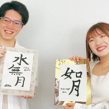 Calligraphy Workshop Two smiling people hold up pieces of paper with Japanese calligraphy. The person on the left wears glasses and a white shirt; the person on the right has short hair and wears a light-colored top. A leafy plant is in the background.