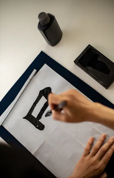 Calligraphy A person practices calligraphy on white paper, using a brush and black ink. An ink bottle and ink stone are placed on the desk nearby. The hand is in the process of writing a bold black character.