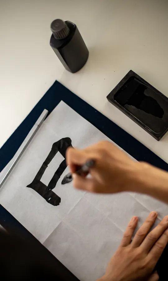 Calligraphy A person practices calligraphy on white paper, using a brush and black ink. An ink bottle and ink stone are placed on the desk nearby. The hand is in the process of writing a bold black character.