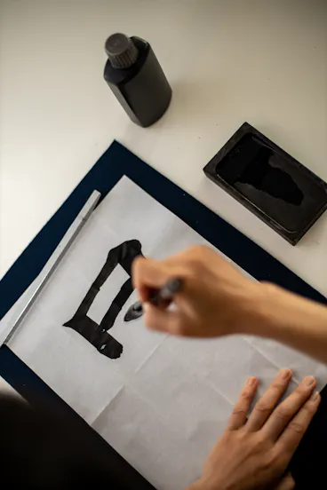 A person practices calligraphy on white paper, using a brush and black ink. An ink bottle and ink stone are placed on the desk nearby. The hand is in the process of writing a bold black character.