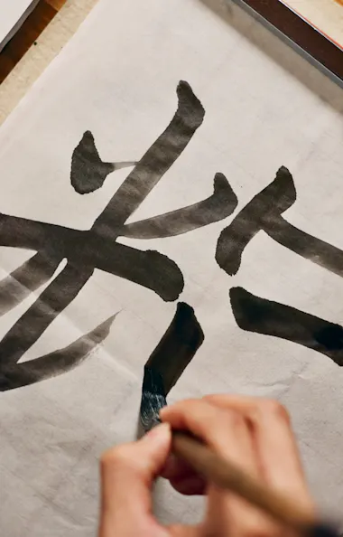 Calligraphy A person practices Japanese calligraphy, painting large black kanji characters on white paper with a brush, on a wooden table.