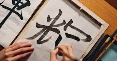 A person practices Japanese calligraphy, painting large black kanji characters on white paper with a brush, on a wooden table.