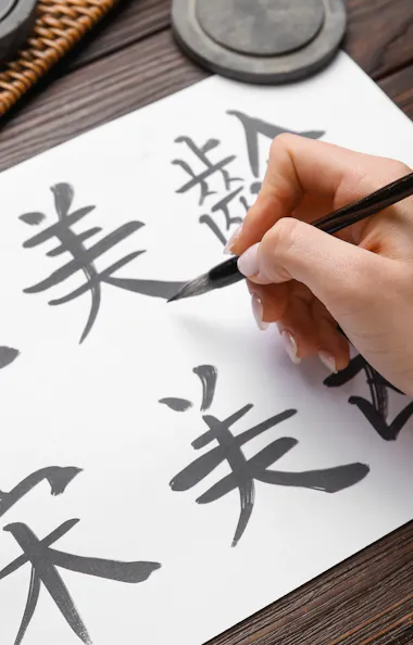 Calligraphy A hand uses a brush to write Chinese calligraphy on white paper. Two cups of tea and ink stones are on a wooden table with a woven mat.