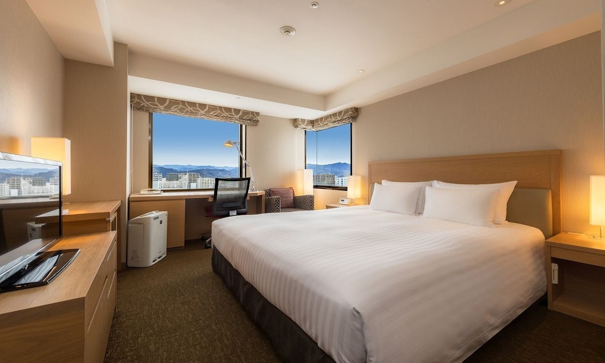 Hotel room featuring a large bed with white linens, a wooden headboard, and bedside lamps. A window reveals a cityscape with mountains in the distance. There's a desk, chair, and large TV. Warm lighting creates a cozy atmosphere.