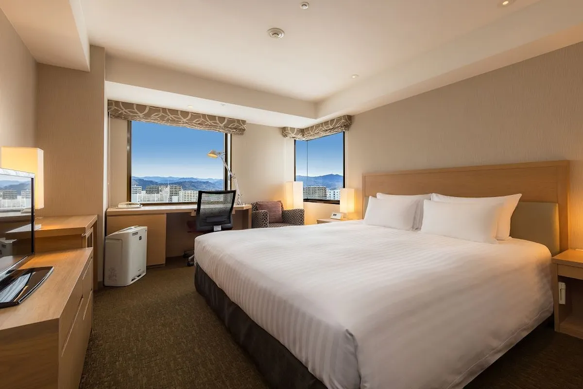 Corner Double room of Hotel Associa Shizuoka Hotel room featuring a large bed with white linens, a wooden headboard, and bedside lamps. A window reveals a cityscape with mountains in the distance. There's a desk, chair, and large TV. Warm lighting creates a cozy atmosphere.