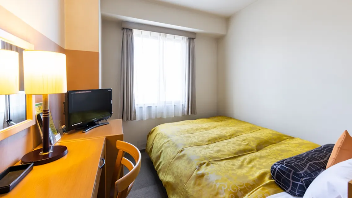A small, bright hotel room with a single bed covered in a yellow bedspread. A window with curtains lets in natural light. A wooden desk with a chair holds a small flat-screen TV and a lamp. The walls are simple and light-colored.