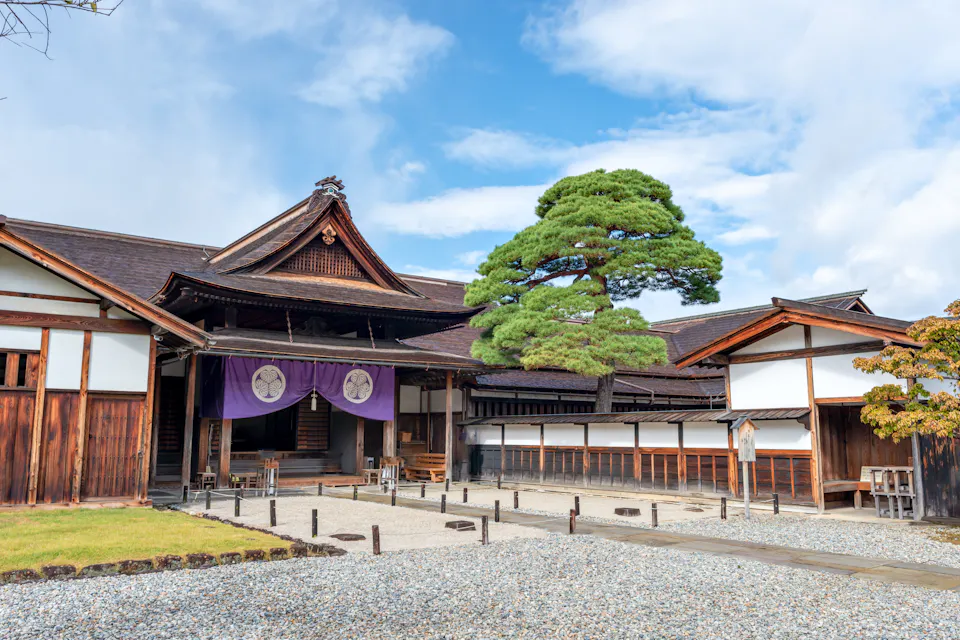 Takayama Jinya Traditional Japanese wooden building with a sloped roof and a decorative purple banner. A pine tree stands prominently in front, with a gravel path leading to the entrance. The sky is partly cloudy.