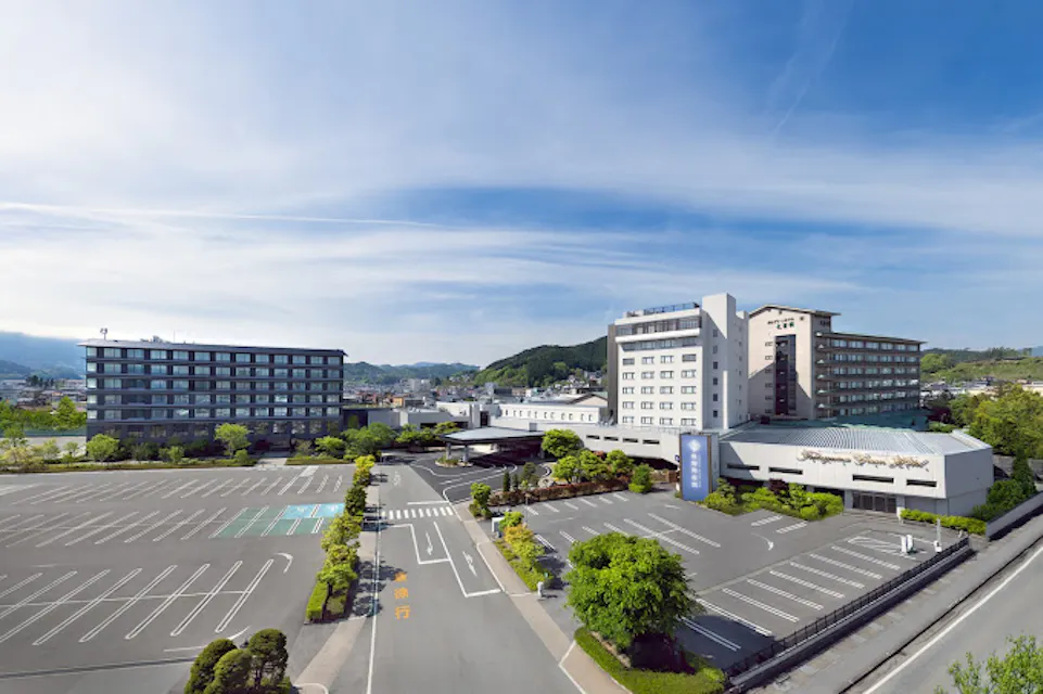 Aerial view of a large hotel complex with multiple buildings and an expansive parking lot. Surrounding greenery and distant hills create a picturesque backdrop under a clear blue sky.