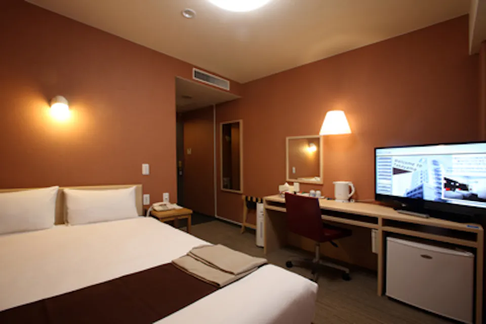 A cozy hotel room with a double bed, white bedding, and brown throw. A desk features a lamp, chair, kettle, and flat-screen TV. A small fridge sits below the desk. Warm lighting and a mirror enhance the space.
