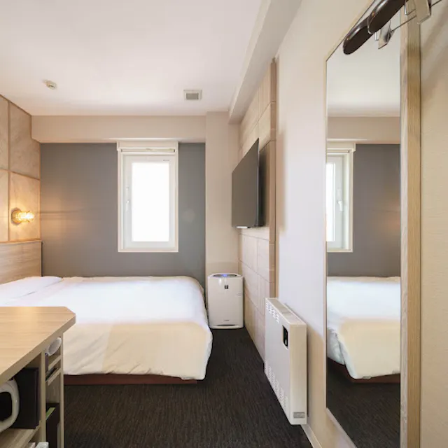 A compact and modern hotel room with a double bed, wall-mounted TV, and a large mirror. The room features a small desk, white walls with a wooden accent, and a window providing natural light.
