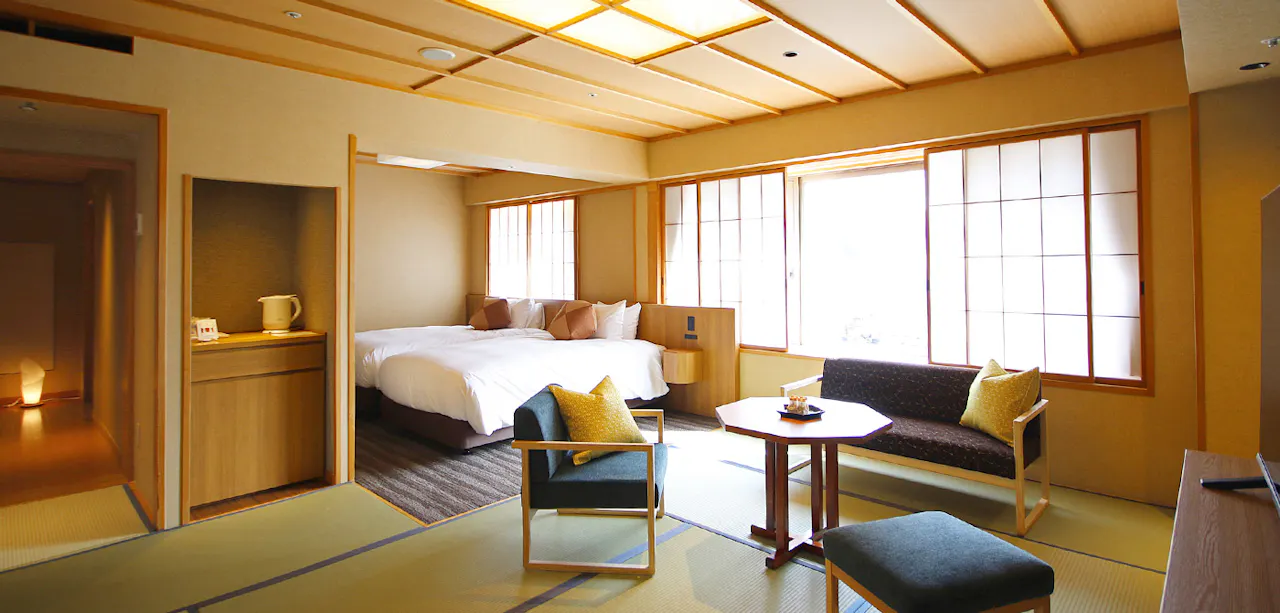 A traditional Japanese-style room with tatami flooring, featuring a bed, two chairs, a small table, and large windows with shoji screens. The room has a minimalistic design with warm wooden accents and soft lighting.