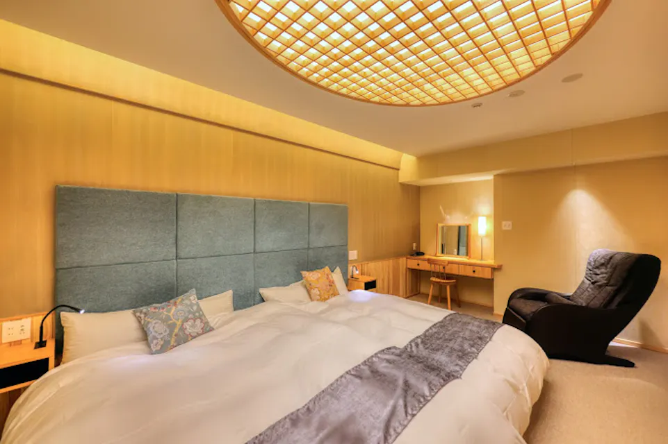 A cozy bedroom features a large bed with a soft blue-gray headboard and patterned pillows. A sleek black chair and a wooden vanity with a mirror are in the corner. The warm lighting creates a relaxing ambiance.