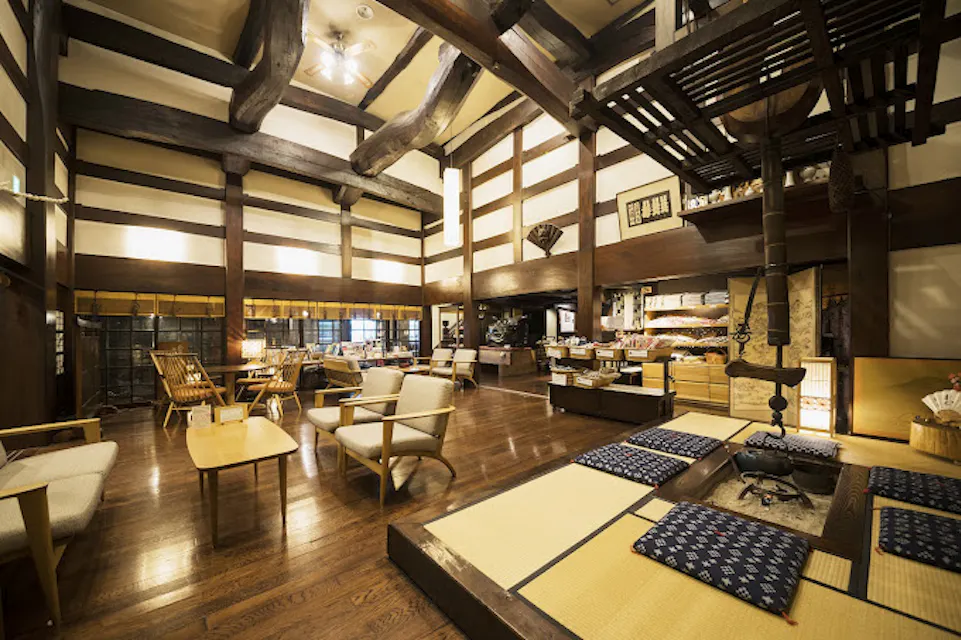 A traditional Japanese room with wooden beams and tatami mats. The space features a low table surrounded by cushions and a sunken hearth. Wooden furniture and soft lighting create a warm, inviting atmosphere.