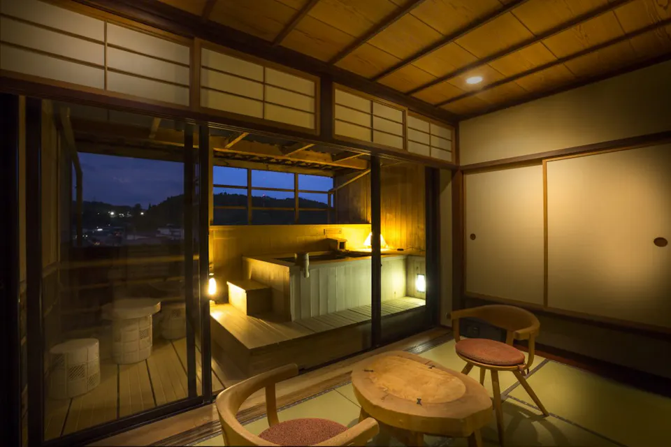 A traditional Japanese room with tatami mats overlooks an open-air bath on a wooden deck. Sliding glass doors separate the spaces. The warm lighting creates a cozy atmosphere, and the night view outside completes the serene setting.