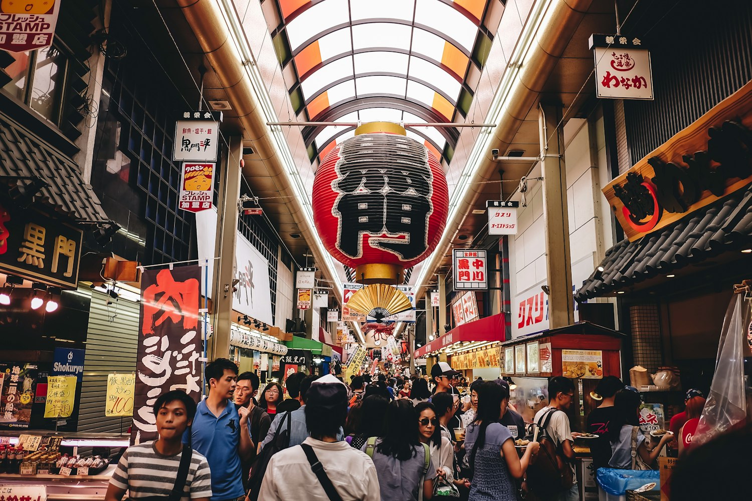A bustling indoor market in Japan with a colorful roof, large red lantern, and a crowd of people shopping. Stalls on either side display various goods and signs featuring Japanese characters.