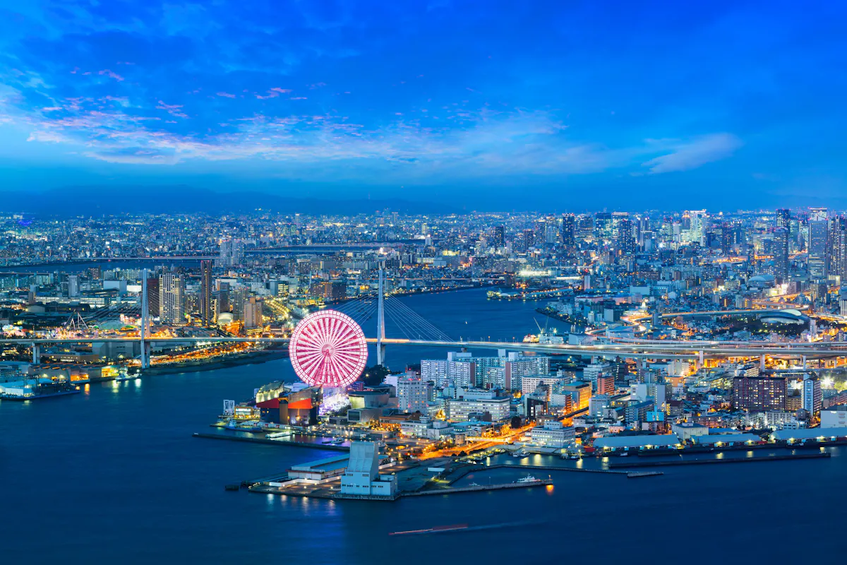 Aerial view of a vibrant cityscape at dusk featuring a large, illuminated Ferris wheel near the waterfront. The sky is a deep blue with scattered clouds, and city lights twinkle across the sprawling urban landscape.