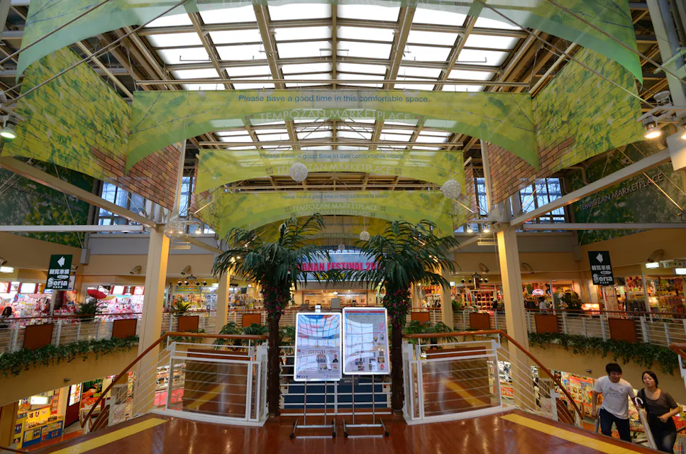 Tempozan Marketplace A vibrant indoor marketplace features high ceilings, a skylight, and colorful stores. Green banners with messages hang overhead. People are walking on the upper level, and a large central plant adds a touch of nature.