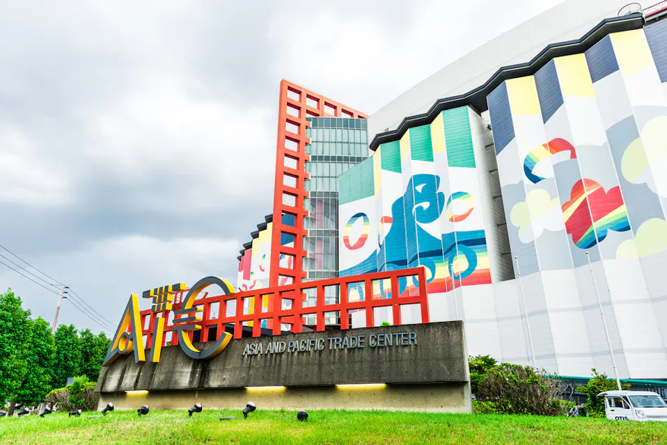 Asia Pacific Trade Center (ATC) The image shows the Asia and Pacific Trade Center with its colorful and modern facade, featuring large abstract murals and red architectural elements. The signage is prominently displayed on a grassy area in the foreground. Cloudy sky in the background.