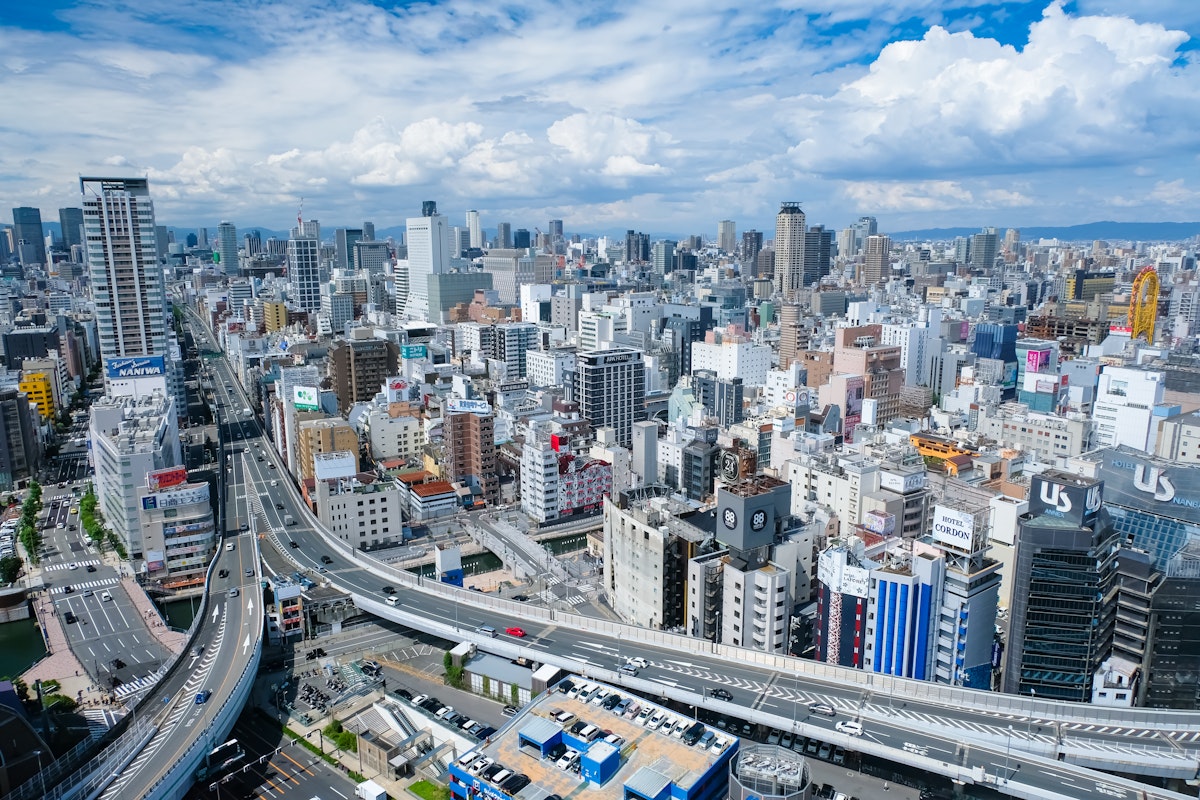 Osaka Minami Area Aerial view of a bustling cityscape featuring numerous high-rise buildings, roads, and highways weaving through the structures. The sky is partly cloudy, adding depth to the vibrant urban environment.