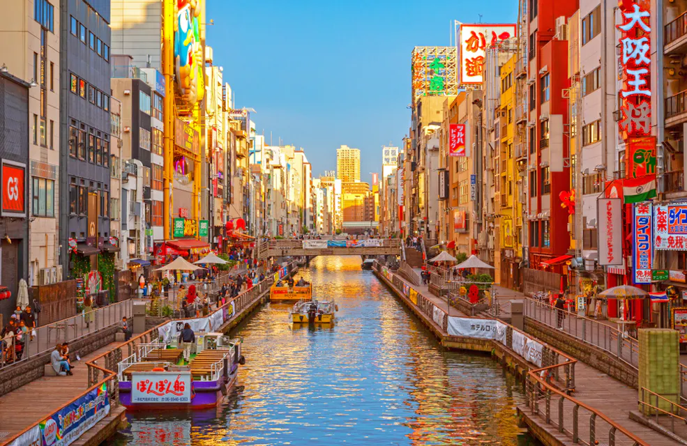 A vibrant canal scene in a city with colorful buildings lining both sides. Boats with people navigate the water, while pedestrians stroll along the pathways. Bright signs in various languages adorn the buildings under a clear, blue sky.