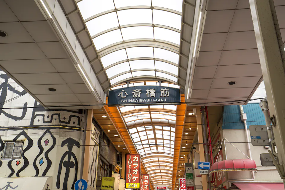 The image shows a covered shopping arcade named Shinsaibashi-Suji in Japan. The area features shops, signs with Japanese characters, and a pedestrian walkway under a canopy of arched glass.