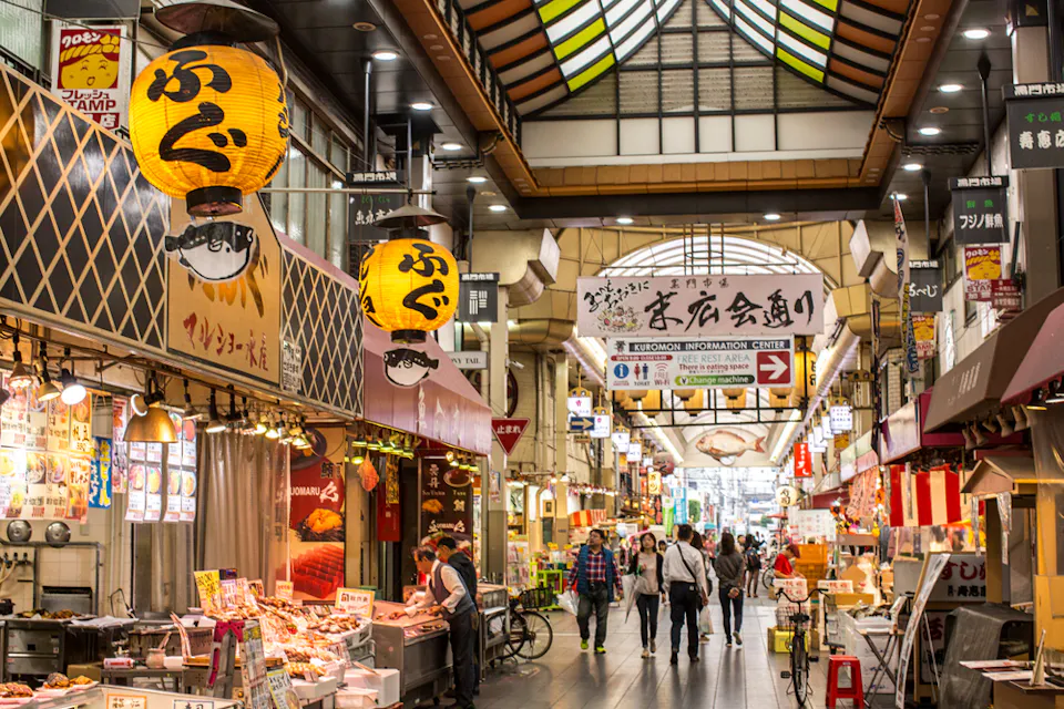 Bustling indoor market street with various stalls selling food and goods. Bright lanterns hang from the ceiling, and signs in Japanese are visible. Shoppers walk between the stalls, creating a lively atmosphere.