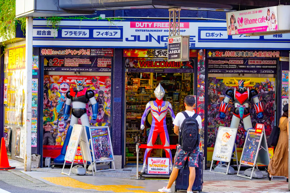 A vibrant storefront features large superhero figures at the entrance. A person with a backpack stands outside, while a colorful array of signs and posters adorn the shopfront. The store appears to sell entertainment and collectibles.