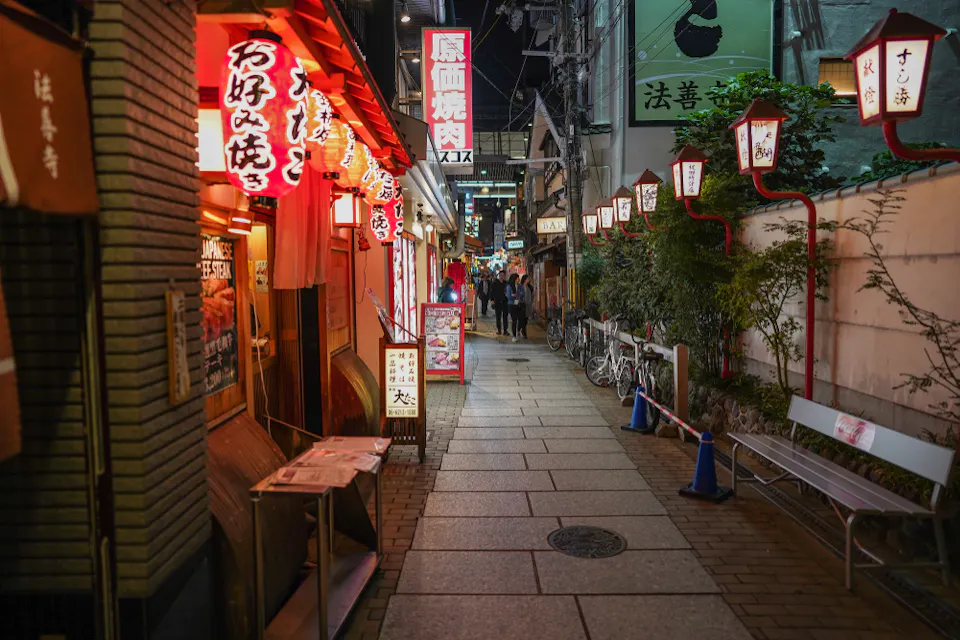 A narrow, charming street in Japan illuminated by red and white lanterns and neon signs. The street is lined with small restaurants, bikes parked on the side, and a couple of people walking in the distance.