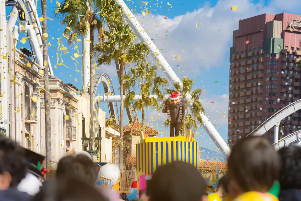 A person in a red Santa hat stands on a striped platform amidst colorful confetti, surrounded by a crowd. Roller coasters and palm trees are visible under a clear blue sky.
