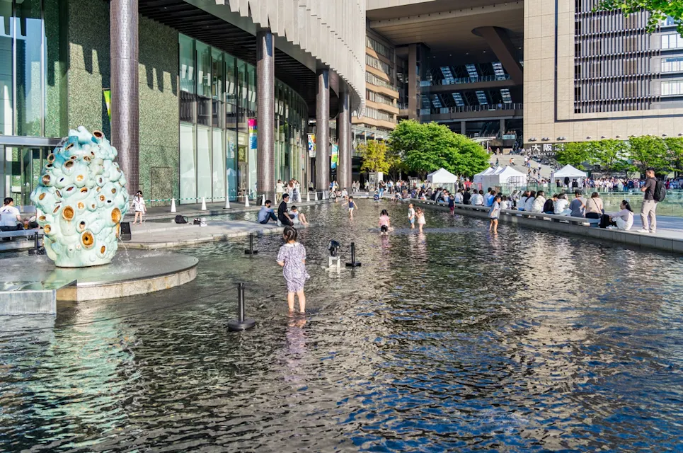 People wading in a shallow, reflective pool surrounded by modern architecture and a sculpture with circular designs. The scene is vibrant with visitors enjoying a sunny day near a building with large glass windows and a leafy area nearby.