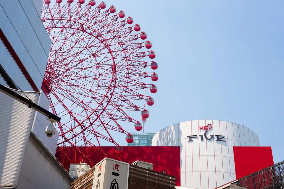 A large red Ferris wheel rises against a clear blue sky, attached to the side of a modern shopping complex with the sign "HEP FIVE." The building has a white exterior with red accents.