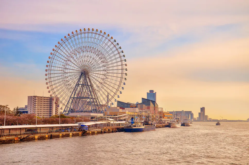 A large Ferris wheel stands near a waterfront with several boats docked along the pier. The sky is softly lit with hues of orange and pink, suggesting sunset. City buildings are visible in the background.