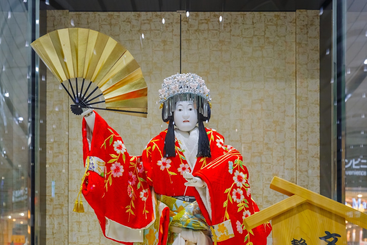 Bunraku A mannequin dressed in a traditional Japanese costume stands in a display. It wears a vibrant red kimono adorned with floral patterns and holds a gold fan. The mannequin's face is painted white.