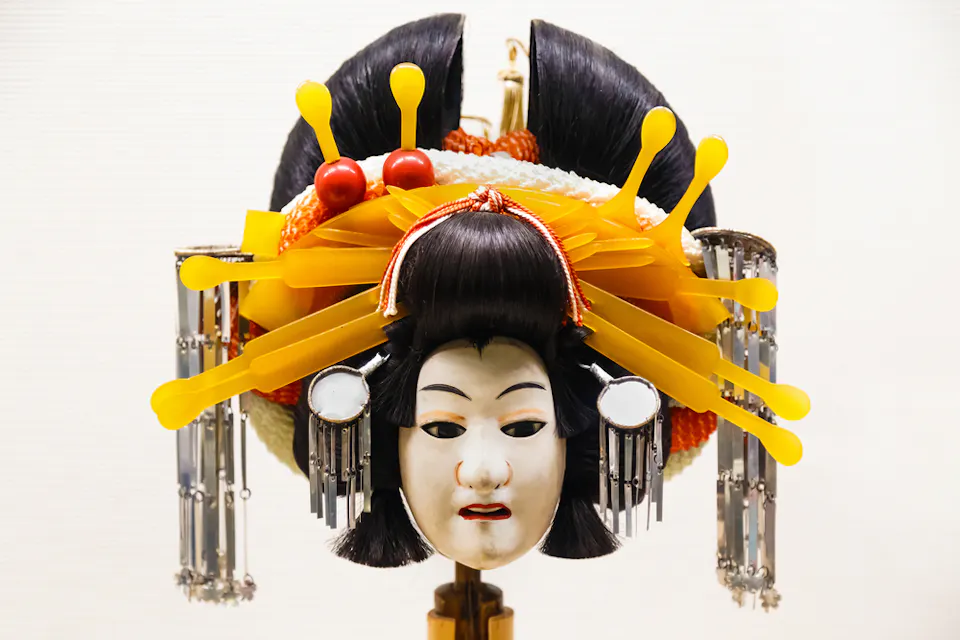 Bunraku A traditional Japanese doll headpiece featuring an ornate hairstyle with decorative yellow and silver hairpins. The face is painted white with red lips and black eyebrows, suggesting a classical theater mask.