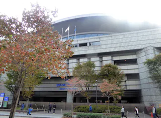 A large, modern building with a curved roof and several floors, surrounded by trees with autumn foliage. People walk on the sidewalk in front of the structure, and flags are seen flying on the roof.