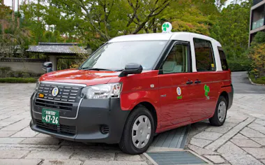 A red and white Japanese taxi with green license plates is parked on a stone street, surrounded by lush greenery and traditional buildings in the background.