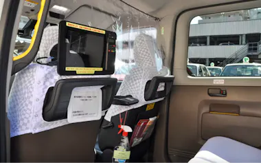 Interior of a taxi with white seat covers, a screen attached to the front seat, clear plastic partition, and a spray bottle for sanitization hanging behind the driver's seat. Daylight is visible outside the window.