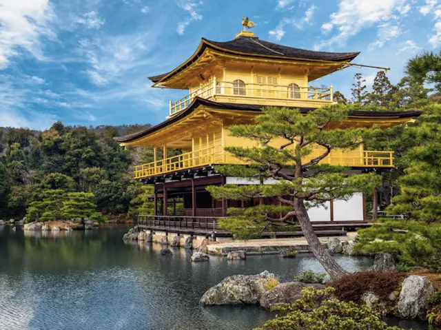 A golden pavilion sits beside a calm pond, surrounded by lush greenery and trees under a partly cloudy blue sky.