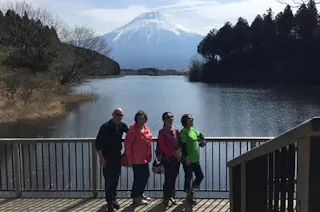 Four people stand on a wooden platform by a lake, with Mount Fuji in the background, surrounded by trees and under a partly cloudy sky.