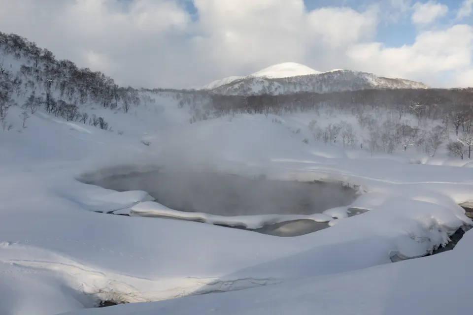A steaming hot spring surrounded by snow-covered hills under a partly cloudy sky. The landscape is blanketed with thick, untouched snow, and a distant mountain peak is visible in the background.