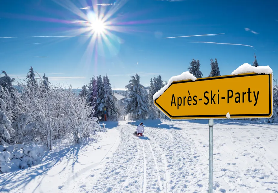 A snowy winter scene with a person sledding down a hill under a bright sun and clear blue sky. Snow-covered trees line the path. A yellow sign reads "Après-Ski-Party" pointing towards the distance.