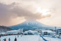 Snow-covered Mount Yotei in the distance with the sun peeking through clouds. In the foreground, a bustling ski resort with skiers and snowboarders on the snow. Buildings and ski slopes are visible under a soft, golden sunset sky.