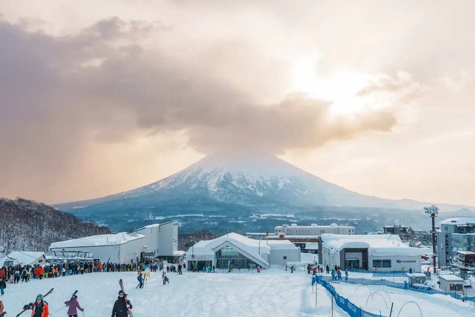 Snow-covered Mount Yotei in the distance with the sun peeking through clouds. In the foreground, a bustling ski resort with skiers and snowboarders on the snow. Buildings and ski slopes are visible under a soft, golden sunset sky.