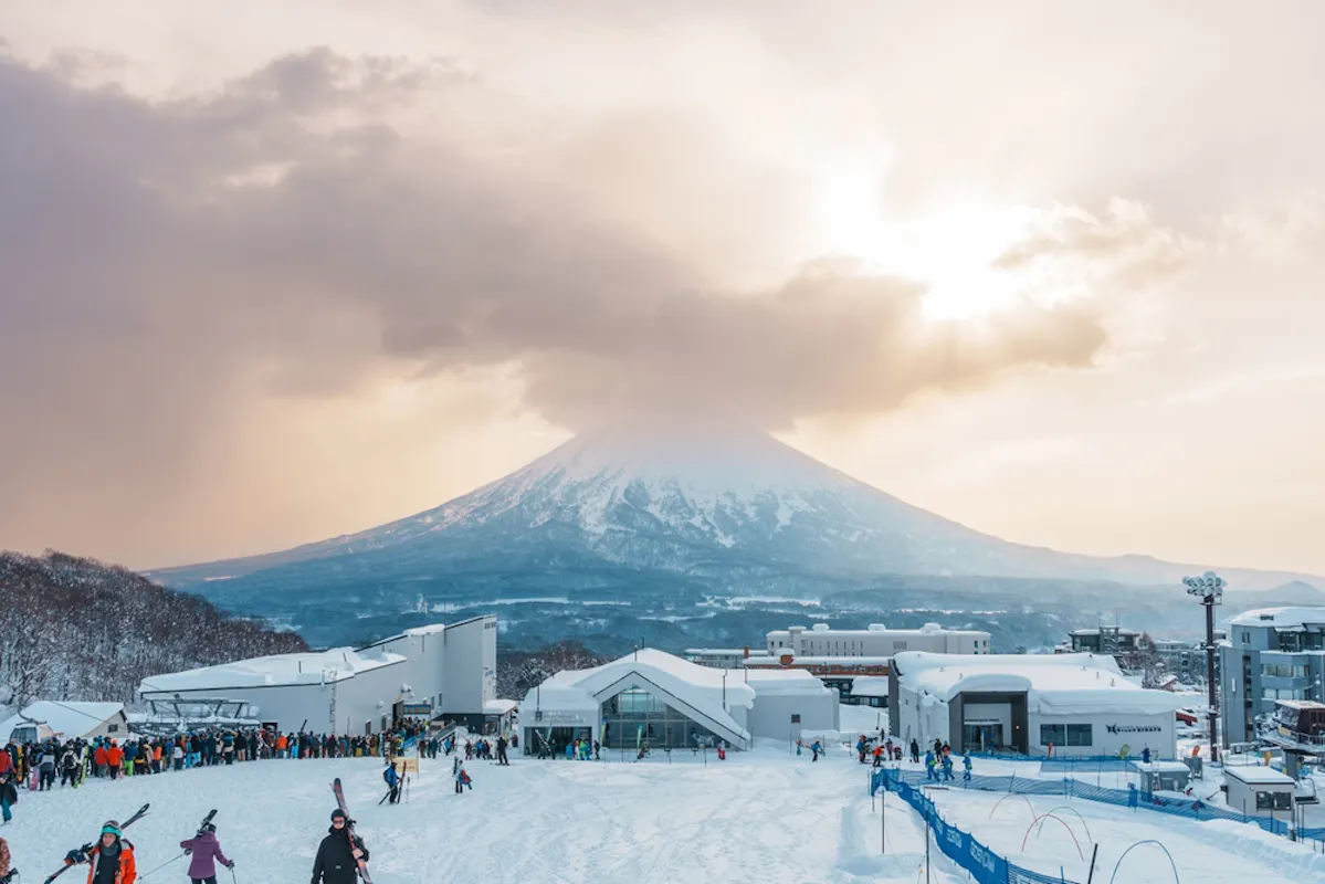 Snow-covered Mount Yotei in the distance with the sun peeking through clouds. In the foreground, a bustling ski resort with skiers and snowboarders on the snow. Buildings and ski slopes are visible under a soft, golden sunset sky.