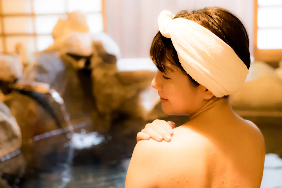 A person with a towel wrapped around their head relaxes in a hot spring, smiling with eyes closed. Water flows gently from a spout into the stone pool. The warm atmosphere is complemented by soft lighting and wooden accents.